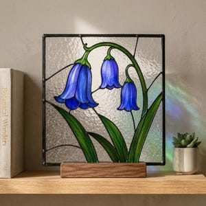 以下が含まれることがあります： 緑の茎と葉を持つ3つの青い鐘形の花が描かれた正方形のステンドグラスパネル。背景は質感のある透明なガラスです。パネルは黒い金属のフレームで囲まれ、長方形の木製ベースに取り付けられています。