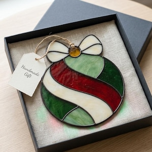May include: A round stained glass ornament featuring diagonal stripes of red, dark green, and cream glass, topped with a cream-colored bow and a small amber center. A white tag with the text "Handmade Gift" is attached by a brown twine string.