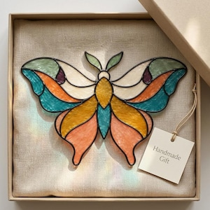 May include: A stained glass moth ornament featuring iridescent wings in shades of teal, orange, yellow, and cream with dark solder lines. The moth is presented in a square box with a beige fabric lining and a white tag labelled Handmade Gift.
