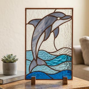 Puede incluir: Un panel rectangular de vidriera que muestra un delfín saltando en tonos azul claro y blanco, sobre un fondo de vidrio transparente texturizado con olas azul oscuro en la parte inferior, sostenido por dos pequeños pies de madera en una superficie.