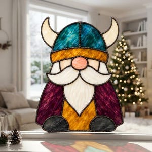 Könnte beinhalten: Ein Buntglas-Sonnenfänger in Form eines Gnoms mit gehörntem Wikingerhelm. Das Stück besteht aus einem blauen Helm, einer pfirsichfarbenen Nase, einem weißen Bart und einem violett-gelben Körper, verbunden durch dunkle Metall-Lötlinien.