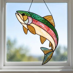May include: A stained glass suncatcher shaped like a leaping trout, featuring iridescent green, pink, and amber glass panels with dark solder lines. The fish is suspended by a thin metal chain, displaying a curved body and detailed fins.