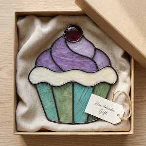 May include: A stained glass cupcake ornament featuring a purple swirled top, a white frosting layer, and a green and teal striped base. A dark red glass cherry sits on top. A small white tag with the text Handmade Gift is attached.