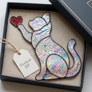 May include: A stained glass cat ornament with an iridescent, textured finish, reaching for a small red heart. The piece is presented in a dark box with a beige fabric lining and includes a white paper tag with the text Handmade Gift attached.