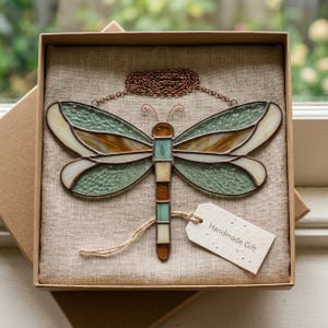 Puede incluir: Un adorno de libélula de vitral con paneles de vidrio verde, ámbar y crema unidos por soldadura de metal oscuro. La libélula tiene una cadena de color cobre para colgar y una pequeña etiqueta de papel blanco con el texto Handmade Gift.