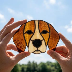 May include: A stained glass suncatcher shaped like a beagle dog head, featuring textured amber and brown glass for the ears and face, a cream-coloured snout, and black glass for the eyes and nose, all held together with dark metal solder lines.
