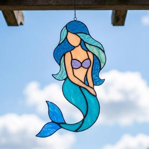 May include: A stained glass suncatcher shaped like a mermaid with flowing blue and teal hair, a peach-colored torso, and a lavender bikini top. The figure features a curved teal tail and is suspended by a thin black metal chain for hanging.