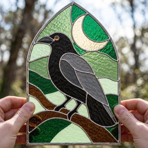 May include: A stained glass panel in a pointed arch shape featuring a black crow perched on a brown branch. The background consists of textured green glass segments and a pale crescent moon. The entire piece is framed in silver metal lead lines.