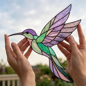 May include: A stained glass hummingbird suncatcher featuring iridescent purple, green, and light pink glass panels held together by dark metal solder lines, depicting the bird in a mid-flight position with wings extended.
