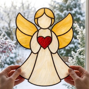 May include: A stained glass angel suncatcher featuring translucent cream-colored glass for the body and head, golden-yellow glass for the wings, and a vibrant red glass heart held at the center, all outlined with dark metal solder lines.