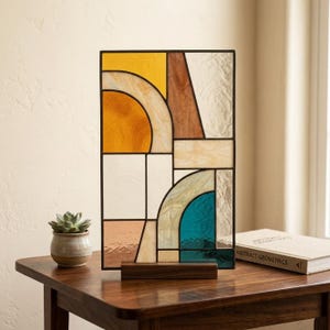 Puede incluir: Un panel rectangular de vidrieras con formas geométricas en amarillo, marrón, crema, melocotón y azul verdoso. Las piezas de vidrio están unidas por líneas metálicas oscuras y montadas sobre una base de madera rectangular para exhibición.