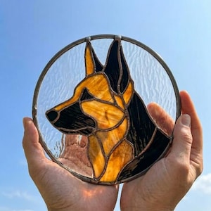 Op de afbeelding: Een ronde glas-in-lood zanger met het profiel van een Duitse herder. Het ontwerp gebruikt amberkleurige en zwarte glasstukken die bij elkaar worden gehouden door donkere metalen soldeerlijnen, tegen een heldere, getextureerde glazen achtergrond.