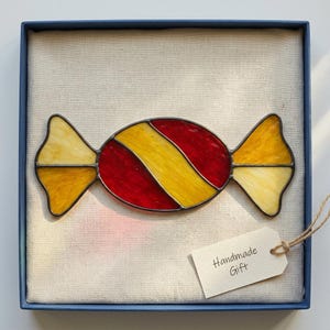 Könnte beinhalten: Ein Buntglas-Bonbon-Ornament mit roten und gelben diagonalen Streifen und dunklen Metalllötstellen, präsentiert in einer blauen Schachtel mit weißem Stofffutter und einem kleinen Papieranhänger mit dem aufgedruckten Text "Handmade Gift".