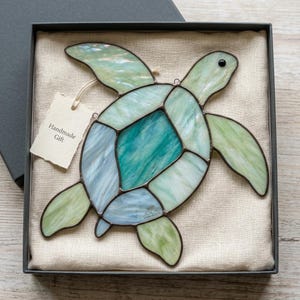 May include: A stained glass sea turtle ornament featuring iridescent light green, teal, and pale blue glass segments joined by dark metal solder lines. A small white paper tag with the printed text Handmade Gift is attached to the turtle.