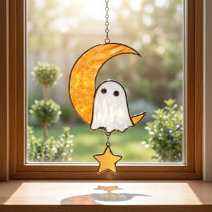 May include: A stained glass suncatcher featuring a crescent moon in mottled orange glass, a white ghost figure with two black circular eyes, and a small yellow star hanging from the bottom, all suspended by a silver metal chain.