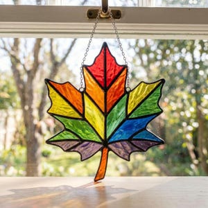 May include: A stained glass maple leaf suncatcher featuring a vibrant rainbow color palette. The leaf is constructed from individual segments of red, orange, yellow, green, blue, and purple glass, held together by dark metal solder lines and suspended by a chain.