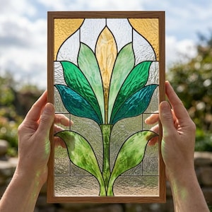 May include: A rectangular stained glass panel framed in light wood, featuring a central floral motif with layered petals in shades of emerald green, teal, and pale yellow, set against a background of clear, textured glass panes.