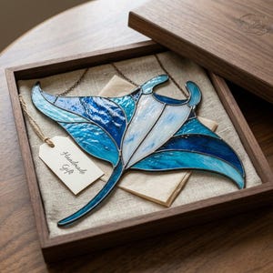 Pode incluir: Um ornamento de vitral em forma de raia manta com peças de vidro em tons de azul profundo, turquesa e branco, unidas por linhas de solda de metal escuro. O ornamento inclui uma pequena etiqueta de papel bege com o texto Handmade Gift.