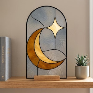 Peut inclure: Un panneau en vitrail arqué représentant un croissant de lune doré et une étoile à quatre branches sur un fond gris translucide et texturé. Les pièces de verre sont assemblées par des lignes de plomb noir sur un socle en bois rectangulaire.