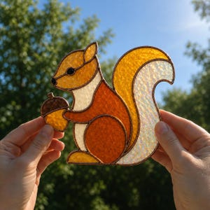 Könnte beinhalten: Ein Buntglas-Suncatcher in Form eines Eichhörnchens, das eine Eichel hält. Das Eichhörnchen besteht aus strukturierten bernsteinfarbenen, orangen und weißen Glasteilen mit Kupferfolienkanten, einem großen, irisierenden Schweif und einem schwarzen Auge.