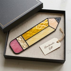 Può includere: Un ornamento in vetro colorato a forma di matita con una gomma rosa, una ghiera dorata testurizzata, un corpo giallo, una sezione in legno color pesca chiaro e una punta grigio scuro. Un'etichetta color crema con la scritta Handmade Gift.