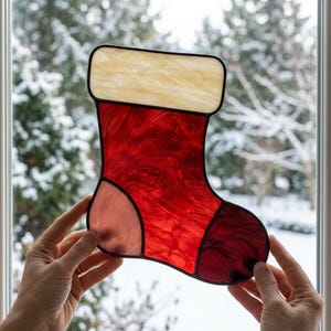 Peut inclure: Une décoration de Noël en vitrail en forme de chaussette, composée d'un revers couleur crème et d'un corps en verre rouge marbré. L'objet est assemblé avec des lignes de soudure en métal sombre soulignant la forme, incluant le talon et la pointe.