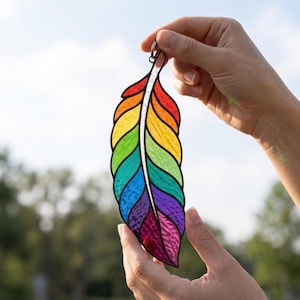Pode incluir: Um ornamento de pena em vitral com um gradiente de cores arco-íris vibrante. A peça é construída a partir de segmentos individuais de vidro vermelho, laranja, amarelo, verde, azul, índigo e violeta, unidos por linhas de solda escuras.