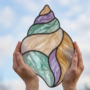 May include: A stained glass seashell suncatcher featuring a spiral design with segments of iridescent cream, soft purple, and seafoam green glass. The individual glass pieces are joined together by dark metal solder lines in a shell shape.