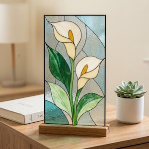 May include: A rectangular stained glass panel featuring two white calla lilies with yellow centers and green leaves. The glass has a textured finish with light blue and clear segments. The panel is held upright by a rectangular wooden base.
