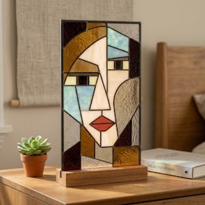 May include: A rectangular stained glass panel featuring an abstract face design with geometric segments in shades of amber, cream, light blue, and dark brown. The piece has a bold red mouth and is mounted on a rectangular light-colored wooden base.