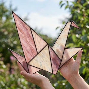 Puede incluir: Una escultura de grulla en vitral construida con paneles geométricos de vidrio veteado en tonos rosa, crema y blanco. Los segmentos de vidrio están unidos por líneas de soldadura de metal oscuro, formando la silueta estilizada de un ave en vuelo.