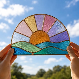 Puede incluir: Un atrapasueños de vidrieras semicircular que presenta un sol ámbar texturizado elevándose sobre colinas onduladas verdes y azules. El cielo está compuesto por paneles radiantes en tonos rosa pálido, amarillo, lavanda y violeta claro, unidos por finos perfiles de latón.