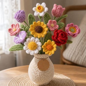 May include: A handmade crochet bouquet of colourful flowers in a beige vase. The bouquet includes sunflowers, daisies, roses, and other flowers in shades of yellow, pink, red, and purple. The vase has a small, blank tag.