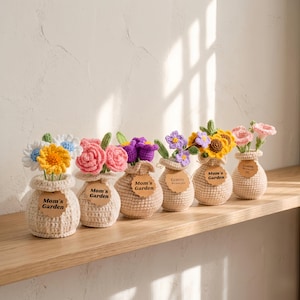 Puede incluir: Seis arreglos florales de ganchillo en pequeños jarrones beige. Cada jarrón contiene un tipo diferente de flor, incluyendo margaritas, rosas y girasoles. Cada jarrón tiene una etiqueta que dice "Mom's Garden" o "Granny We love you".