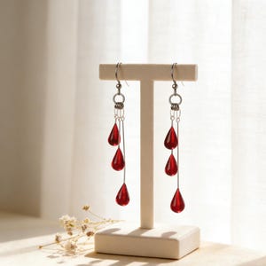 May include: Dangling earrings with three red teardrop-shaped beads suspended from silver-colored hooks. The earrings are displayed on a white stand against a soft, neutral background, highlighting their vibrant color and design.