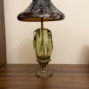 May include: A table lamp featuring a wide, dark brown glass shade with white speckled patterns. The base is made of a translucent olive green glass with an organic, fluid shape, supported by a decorative, ornate brass-colored metal stand and base.