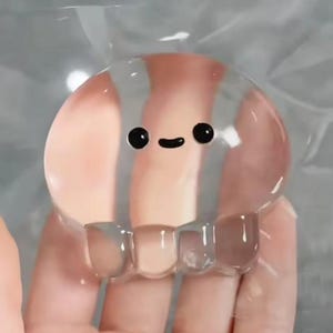 May include: A clear, jelly-like, rounded object shaped like a jellyfish with a smooth, translucent surface. It features a simple, hand-painted black face consisting of two small circular eyes and a curved mouth centred on the upper portion.