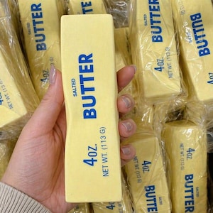 May include: A hand holding a 4 oz. stick of salted butter, with the word "BUTTER" in blue. Other wrapped butter sticks are visible in the background. The net weight is 113 g.