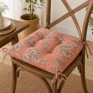 May include: A square seat cushion in a coral orange hue featuring a detailed floral pattern in muted tones of beige and grey. The cushion is quilted with vertical stitching and includes four fabric ties at the corners to secure it to a chair.