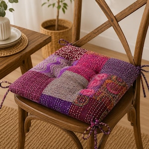May include: A square seat cushion featuring a patchwork design of various purple, magenta, and pink fabrics with intricate white kantha stitching. The cushion includes fabric ties at the corners for securing it to a wooden chair seat.