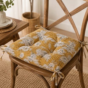 May include: A square chair cushion featuring a mustard yellow and white floral pattern with a textured, grid-like fabric surface. The cushion includes four corner ties for securing it to a wooden chair and has a tufted design with visible stitching.