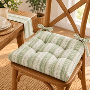 Puede incluir: Un cojín de silla cuadrado con un patrón de rayas verde claro y blanco con pequeños motivos de diamantes. El cojín está acolchado en cuatro secciones e incluye dos lazos de tela en las esquinas traseras para fijarlo a la silla de madera.