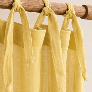 Può includere: Un pannello per tenda giallo chiaro realizzato in tessuto testurizzato semitrasparente. Il bordo superiore presenta lunghi lacci in tessuto annodati saldamente attorno a un bastone per tende cilindrico in legno, creando un aspetto drappeggiato.