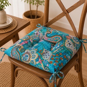 May include: A square chair cushion featuring a vibrant turquoise base with a multi-colored paisley and floral pattern. The quilted fabric has a soft texture and includes four fabric ties at the corners for securing the cushion to a wooden chair.