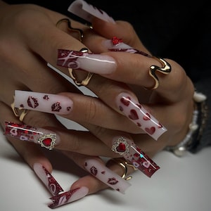 May include: Long, square-shaped acrylic nails with a clear base and various designs. Some nails feature red hearts, lips, and glitter accents. Gold rings adorn the fingers. A heart-shaped jewel is on one nail.