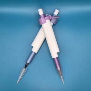May include: Two white and purple pens with clear, cone-shaped tips are crossed against a blue background. The pens have a cylindrical design with a purple band and a purple and blue gradient on the lower portion.