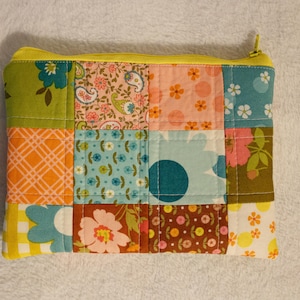 May include: A patchwork zippered pouch with a yellow zipper and trim. The pouch is made of various fabric squares in floral and geometric patterns, including orange, green, pink, and blue. The pouch is quilted.