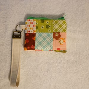 May include: A small, patchwork fabric wallet with a green zipper and a cream-colored wrist strap. The wallet is made of squares of fabric in various patterns and colors, including floral and geometric designs. The wrist strap is attached with a gold-colored metal clasp.