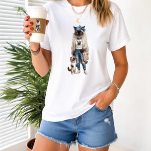 Może przedstawiać: Biała koszulka z grafiką kota w dżinsach, niebieskiej opasce i torebce, z mniejszym kotem. Koszulka jest połączona z dżinsowymi szortami. W dłoni trzymany jest kubek do kawy z grafiką kota.