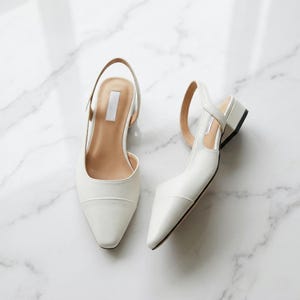 Puede incluir: Un par de zapatos de tacón blancos con punta y tacón de bloque pequeño. Los zapatos tienen una correa que rodea el tobillo y una plantilla de color neutro. Los zapatos están hechos de un material liso.